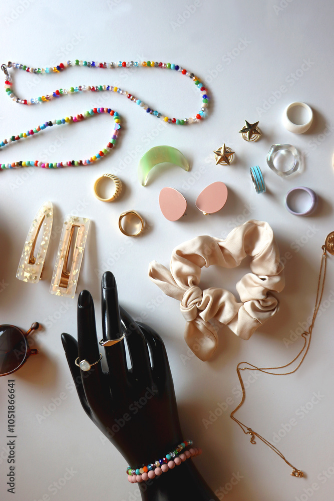 Hand figurine, sunglasses, gold jewelry, colorful bead jewelry, statement rings and various hair accessories on the white table. Flat lay.