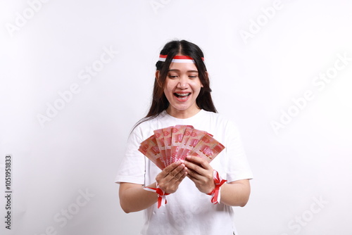 Excited Asian woman wearing red white flag ribbon, holding indonesia banknotes rupiah money, isolated on a white background, celebrate Indonesia independence day photo