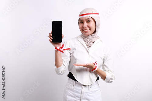 Smiling young Asian Muslim woman showing mobile phone blank screen with her hand isolated over white background. Celebrate Indonesian independence day on 17 August photo