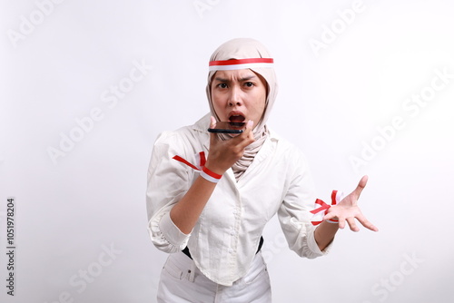 Angry young Asian Muslim woman wearing a hijab shouting loudly at mobile phone isolated over white background. Celebrate Indonesian independence day on 17 August photo