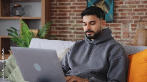 Careless indian male keeping modern laptop on legs and typing while looking at display with toothy smile. Bearded man with ear piercing testing new computer and checking available functions.