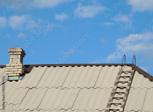 chimney brick pipe and wooden ladder on gray corrugated slate roof of private rural house under clear blue sky, top of rustic building with stairs and kitchen pipe for smoke outlet as country texture photo