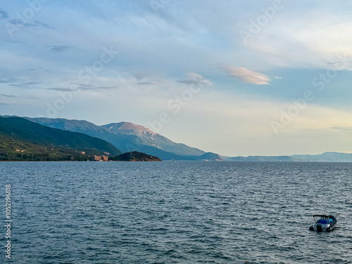 Small boats floating on calm Lake Ohrid in North Macedonia. Rolling hills covered in lush green vegetation. Tranquil lakeside scene. Summer travel destination in the Balkans. Boat tour tourism