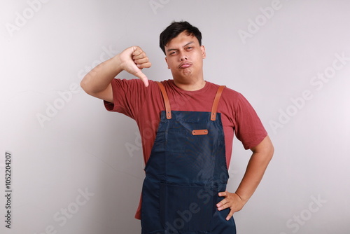 Moody unsatisfied asian man wearing blue apron, disliking bad restaurant food smirking displeased, grimacing disappointed show thumb down, shaking head negative judgement, stand white background