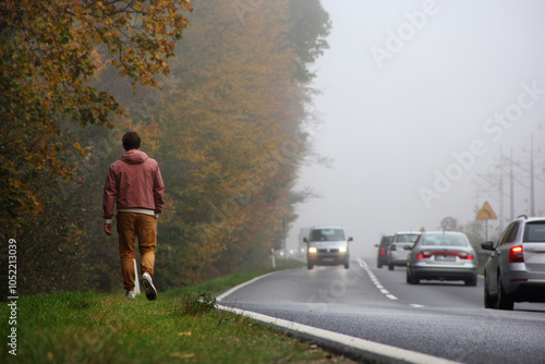 A young man in pants and a jacket walks along the autumn roadside along an autumn forest and an intercity highway along which cars are driving with their headlights on, on a foggy day photo