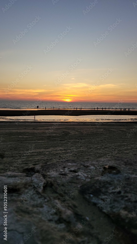 Fototapeta premium This is a beautiful sunset photograph taken on a beach. The sun is low on the horizon, casting warm orange and yellow hues across the sky and reflecting on the water. The scene includes several people