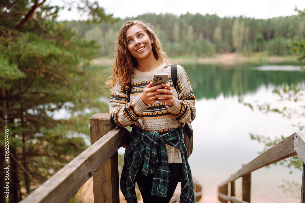 A young woman in a cozy sweater walking near a beautiful lake, smiling and using her phone. Adventure, nature, blogging. A young woman in a cozy sweater walking near a beautiful lake, smiling and using her phone. Adventure, nature, blogging.