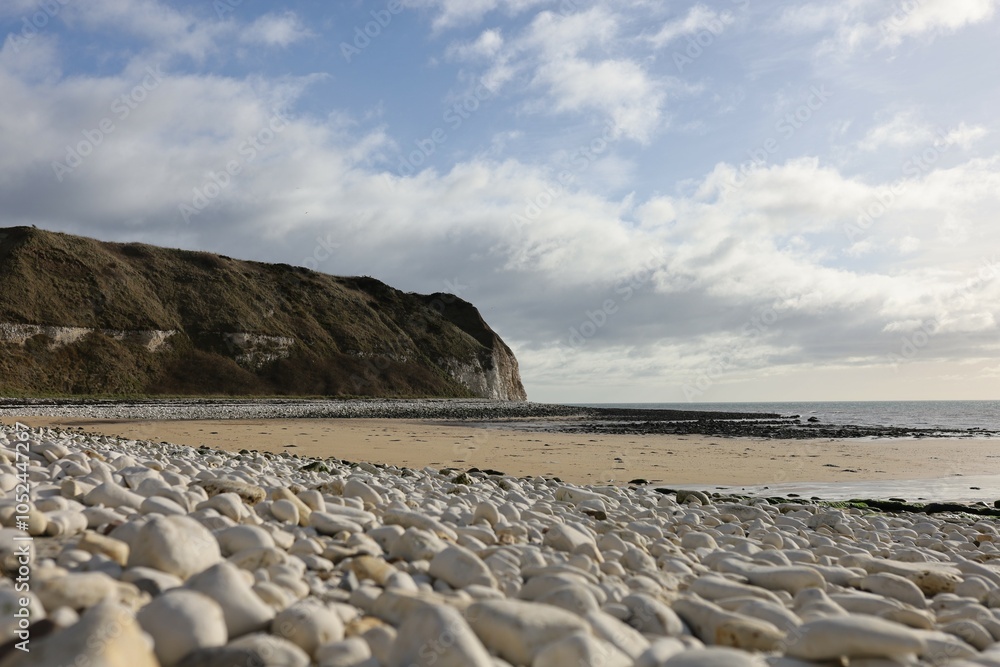 Seaside landscape with large white stones lying on the shore in the foreground. Behind them,a sandy beach leads into a calm sea. In the distance,there is a tall cliff covered with grass and vegetation