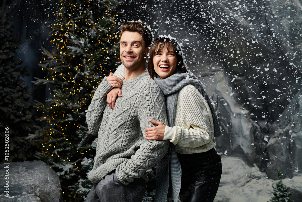 A couple bundled in cozy grey sweaters enjoys a festive winter moment near snow-covered fir trees. A couple bundled in cozy grey sweaters enjoys a festive winter moment near snow-covered fir trees.