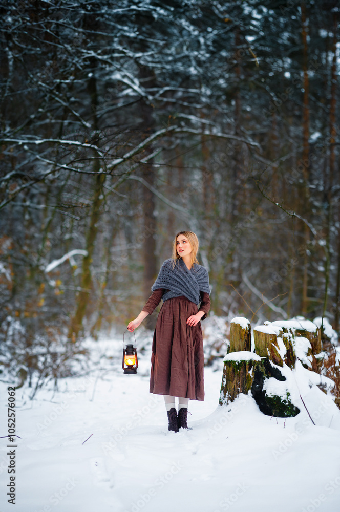 Naklejka premium Portrait of beautiful blonde girl in medieval dress and woolen scarf in winter snowy forest. Young worker in rural scene with flashlight.Colorful fairytale art work.