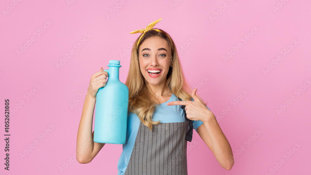 Smiling housewife points finger at fabric softener, on pink background, empty space Smiling housewife points finger at fabric softener, on pink background, empty space