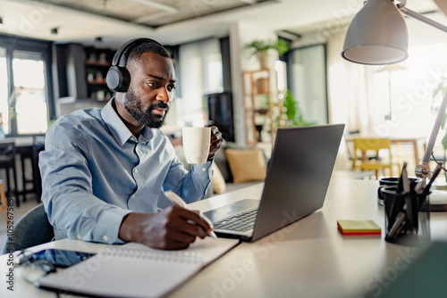 African-American man wearing headphones, taking notes while working on a laptop from home, focused and engaged in a modern workspace.