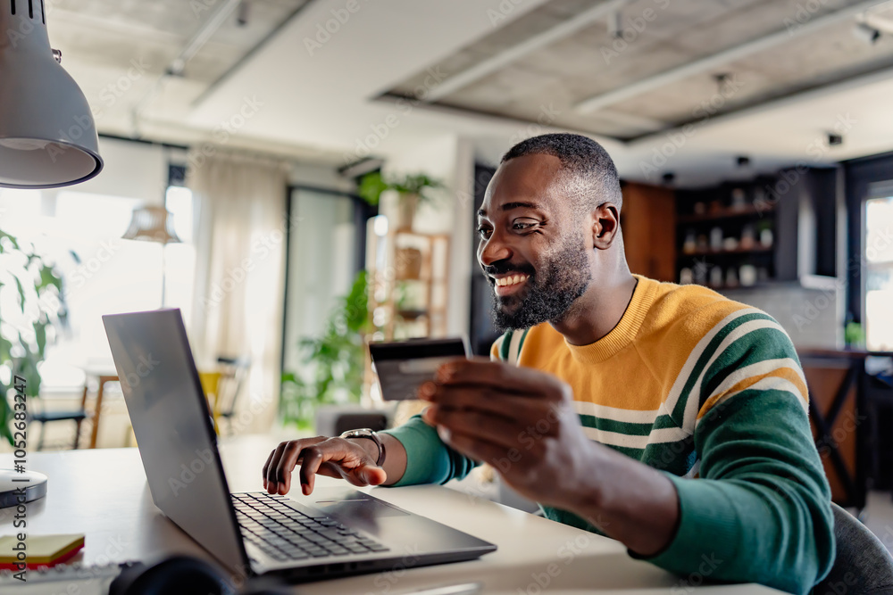 Smiling young African-American man using a debit card for online shopping or banking on his laptop at home. He is seated in a cozy, modern workspace, appearing engaged and productive. Smiling young African-American man using a debit card for online shopping or banking on his laptop at home. He is seated in a cozy, modern workspace, appearing engaged and productive.