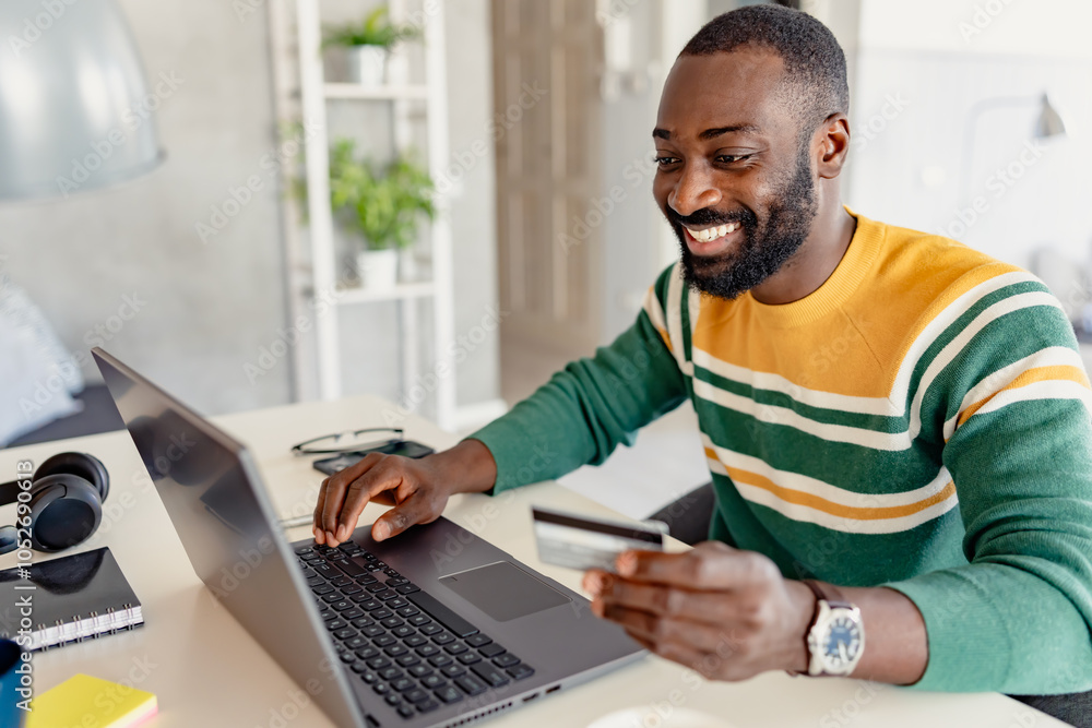 Smiling young African-American man using a debit card for online shopping or banking on his laptop at home. He is seated in a cozy, modern workspace, appearing engaged and productive. Smiling young African-American man using a debit card for online shopping or banking on his laptop at home. He is seated in a cozy, modern workspace, appearing engaged and productive.