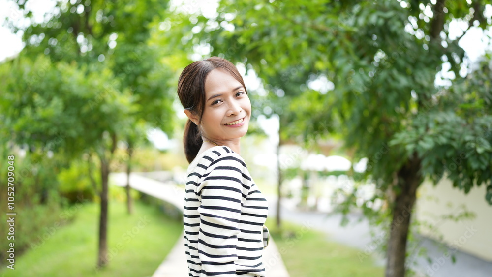 young woman smiles joyfully while standing on pathway surrounded by lush greenery. vibrant trees and soft sunlight create cheerful atmosphere young woman smiles joyfully while standing on pathway surrounded by lush greenery. vibrant trees and soft sunlight create cheerful atmosphere