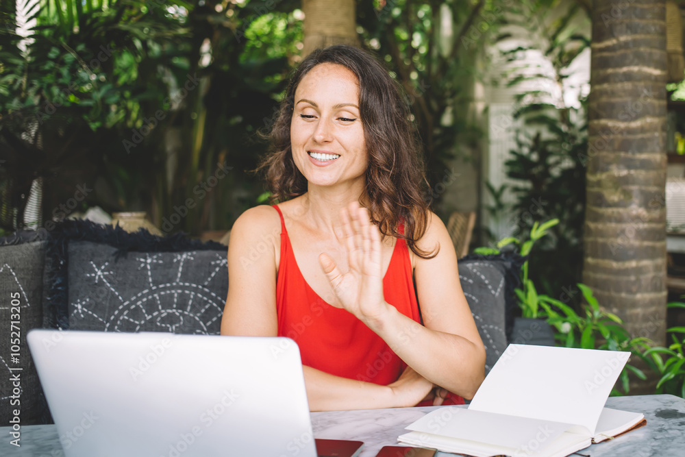 Smiling woman in red top waving during a video call on her laptop in a vibrant outdoor workspace. She embodies a friendly, professional digital nomad connecting remotely, surrounded by greenery Smiling woman in red top waving during a video call on her laptop in a vibrant outdoor workspace. She embodies a friendly, professional digital nomad connecting remotely, surrounded by greenery