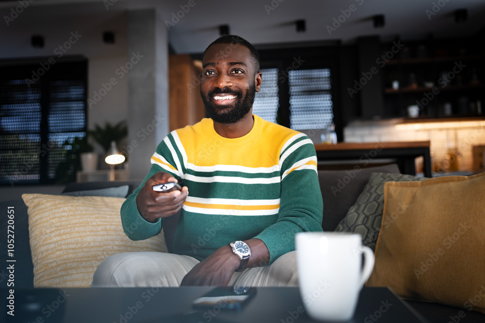 Smiling African-American man sitting on a couch, holding a remote control and watching TV in a cozy living room with a warm atmosphere Smiling African-American man sitting on a couch, holding a remote control and watching TV in a cozy living room with a warm atmosphere
