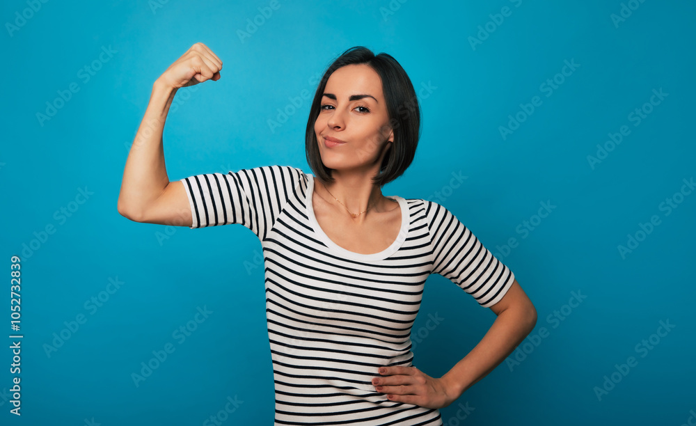 A young woman in a striped shirt flexes her arm with a confident expression against a blue background. Her pose conveys strength, empowerment, and self-confidence, embodying a
