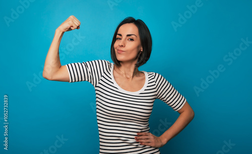 Obraz na płótnie A young woman in a striped shirt flexes her arm with a confident expression against a blue background. Her pose conveys strength, empowerment, and self-confidence, embodying a