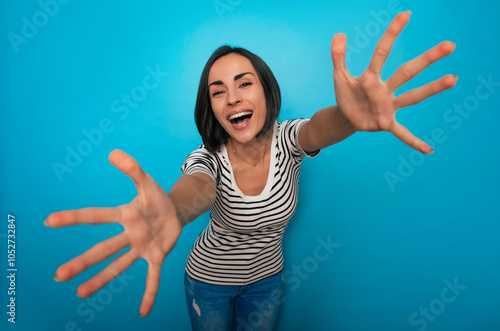 A joyful woman with short dark hair and a striped shirt reaches her arms forward toward the camera, smiling brightly, set against a blue background, conveying warmth, excitement, and friendliness. photo