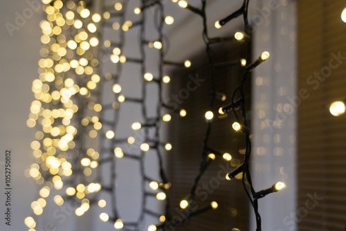 Warmly flickering garland lights against golden blinds, seen from the side, bring a cozy, holiday glow. The distant bulbs create a gentle bokeh effect, adding depth and festive sparkle to the photo.