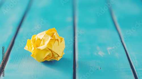 A crumpled yellow paper ball rests on a wooden surface, creating a vibrant contrast against the mellow tones of the backdrop. photo