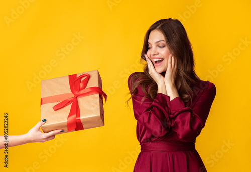 Surprise For You. Excited young lady in evening dress getting unexpected gift box, touching cheeks in joy, standing over colorful background, free space