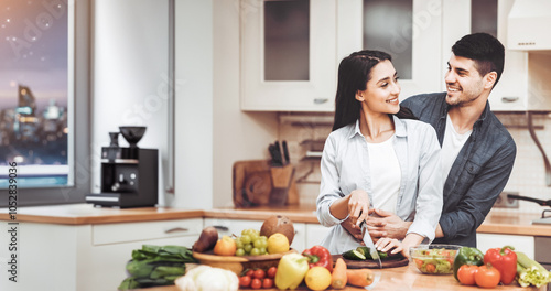 Happy Loving Family. Young lady cooking salad at kitchen, guy embracing her from the back, copy space