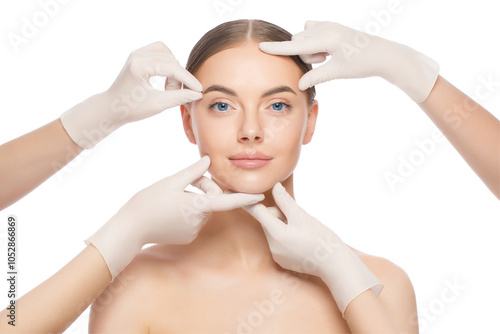 Close-up portrait of young female standing with naked skin, face is touched by doctors in gloves, preparing her for plastic surgery procedures