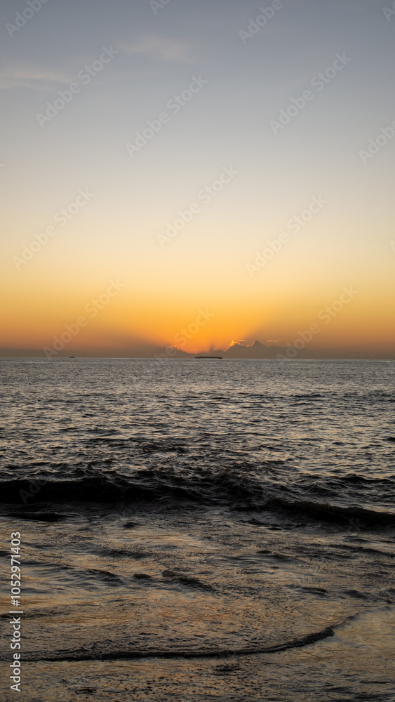 Obraz premium A calm seascape displays the bright colors of a tropical sunset, with waves gently lapping the shore and distant clouds illuminated by the last rays of the sun, creating a peaceful beach scene.