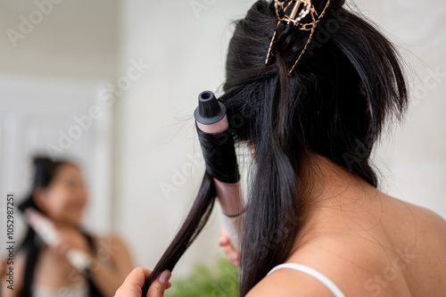 A close-up of an Asian woman with long, natural black hair curling her hair with a curling iron.