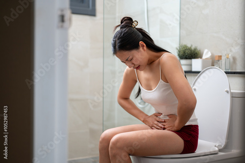 An Asian woman in pajamas sitting on a toilet, gently touching her stomach, severe stomach pain.