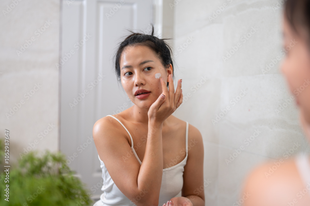 A natural Asian woman is applying facial cream in front of the mirror in the bathroom after shower. A natural Asian woman is applying facial cream in front of the mirror in the bathroom after shower.
