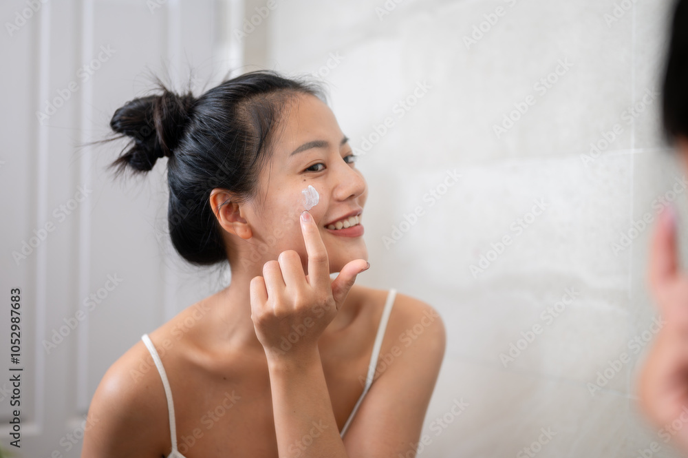 An attractive Asian woman is smiling at the mirror while applying facial cream to her face. An attractive Asian woman is smiling at the mirror while applying facial cream to her face.