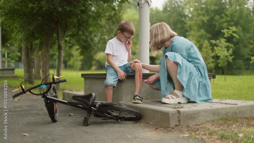 A concerned mother kneels beside her injured child, cleaning a bloody knee wound with a tissue, as the boy chases flies from the wound A concerned mother kneels beside her injured child, cleaning a bloody knee wound with a tissue, as the boy chases flies from the wound