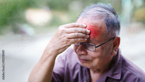 Elderly man with glasses clutching his forehead in pain, showing a red bruise on his head, indicating an injury. Outdoor setting. photo