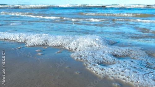 Footage captures sea foam slowly moving along with the water as waves reach the sandy shore at Vääna Jõesuu beach. Foaming waves and clear blue sky are in the background during beautiful sunny day. photo