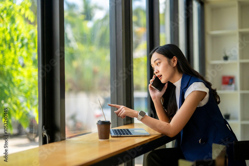 A woman is talking on her cell phone while pointing to something on her laptop