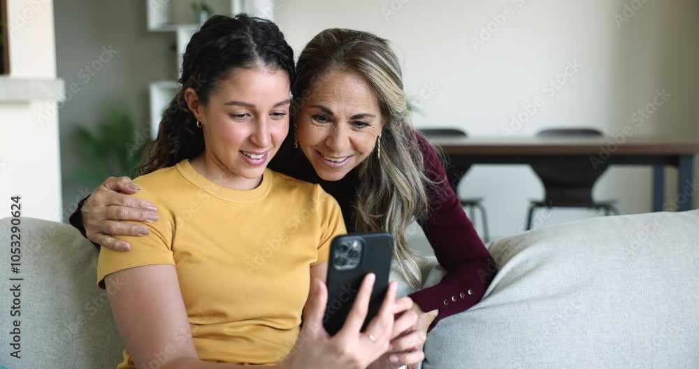 Positive young adult Latin daughter and mature mom watching online video content on mobile phone together, getting cheerful, laughing, enjoying Internet communication, technology at home Positive young adult Latin daughter and mature mom watching online video content on mobile phone together, getting cheerful, laughing, enjoying Internet communication, technology at home