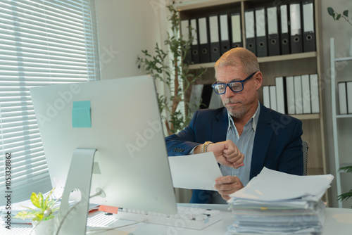 Senior manager looking at his watch while reviewing a document, sitting at his desk in a bright office, possibly waiting for a meeting or appointment photo