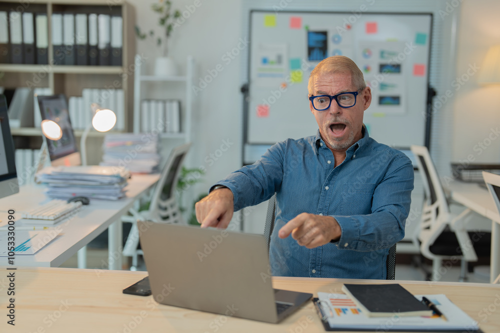 Senior manager looking surprised while pointing at his laptop screen, sitting at his desk in a modern office, surrounded by paperwork and technology Senior manager looking surprised while pointing at his laptop screen, sitting at his desk in a modern office, surrounded by paperwork and technology