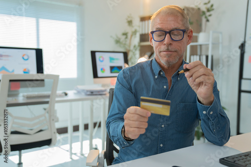 Senior businessman holding credit card and pen, inputting information for online shopping, paying bills or registering for membership in modern office with computers showing charts