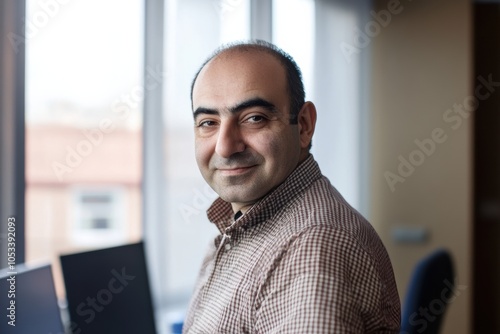 Professional portrait of a smiling man from Azerbaijan, dressed in a brown checkered shirt, set against a modern office background.