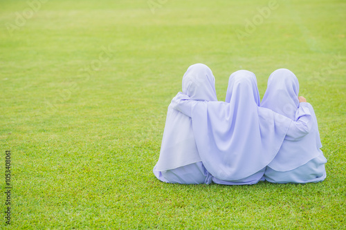 Three girls wearing white hijabs sit hugging each other on a green grass background.