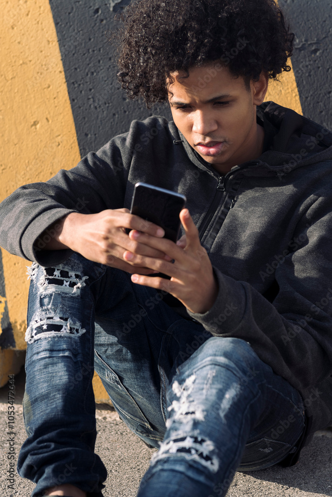 serious african american male teenager in ripped jeans using smartphone while sitting near striped wall on street, concept of technology of communication and young lifestyle serious african american male teenager in ripped jeans using smartphone while sitting near striped wall on street, concept of technology of communication and young lifestyle