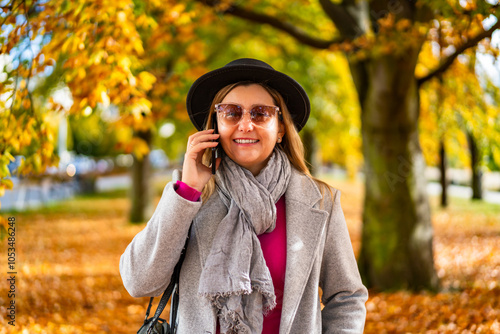 Portrait of smiling middle aged woman wearing casual clothes, black brimmed hat and glasses walking, talking on phone against alley with deciduous trees in park in background on sunny autumn day. 