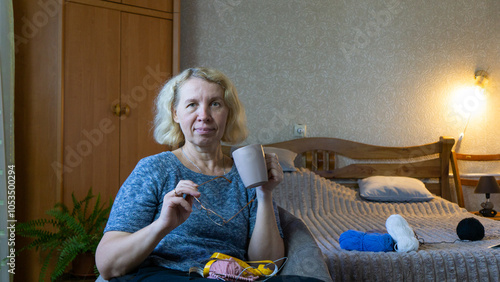 A middle-aged woman enjoys a drink from a cup, holding reading glasses in her hand. Cozy home atmosphere. Skein of yarn lie on bed