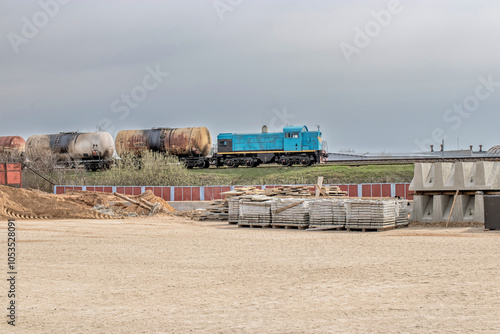 A blue locomotive pulls freight cars past industrial debris at a construction site under overcast skies in the daytime photo