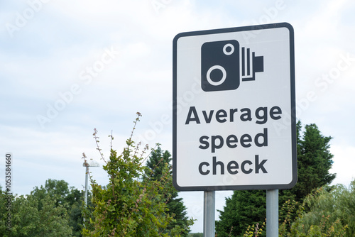 Large roadside sign notifying motorists, that the stretch of highway ahead is an Average Speed Check zone. Low view of sign against a clear sky with green bushes nearby.
