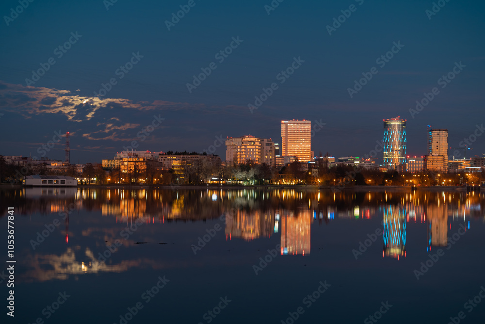 Obraz premium Amazing urban landscape with tall buildings illuminated at night and reflected in the water. Moonrise above the skyscrapers of Bucharest, the capital of Romania